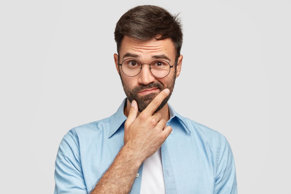 Na imagem vemos um rapaz com cara de dúvida, ele está com uma camisa social, e leva sua mão ao queixo. Ele representa o leitor que irá descobrir as12 perguntas essenciais para escolher um software de gestão de processos