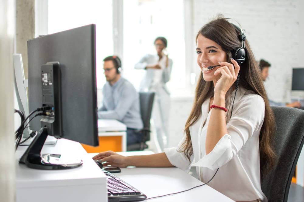 na imagem vemos uma moça com headset, ela está sorrindo. Atrás dela, há outras pessoas trabalhando no escritório. Ela representa o suporte técnico na implantação de ferramentas de gestão de processos
